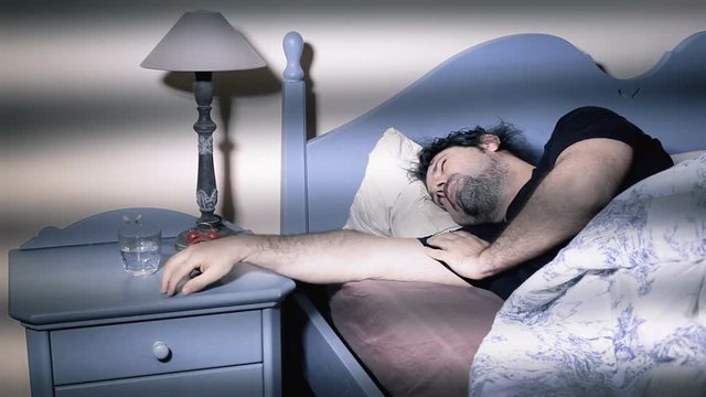 A tired sleeping man in bed in the early morning picks up the ringing alarm clock and puts it into a glass of water to make it stop.
