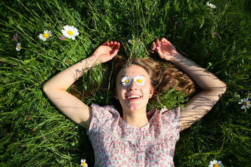 Smiling happy relax young woman lying on fresh green grass in daisies in eyes, flowers, outdoor, summer background, top view, rural