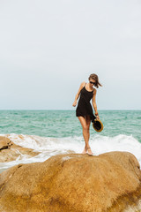 Beautiful sporty woman in little black dress and summer straw hat dance on rock with waves under. Beauty cute girl on a tropical beach sea ocean shore with large stones. Outdoor summer lifestyle.