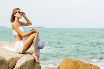 Fototapeta na wymiar Beautiful woman in white skirt, top and summer straw hat sit on edge of rock. She correct sunglasses. Beauty cute girl on a tropical beach sea ocean shore with large stones. Outdoor summer lifestyle.