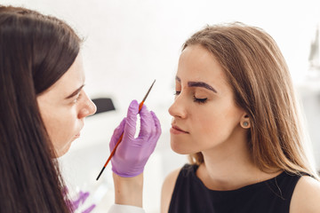Portrait of professional cosmetologist in purple gloves making permanent eyebrows for woman, using brush. Female client with closed eyes smiling and enjoying in beauty salon.
