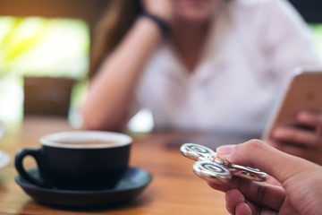 A hand holding and playing metal silver color fidget spinner with coffee cups and people in background