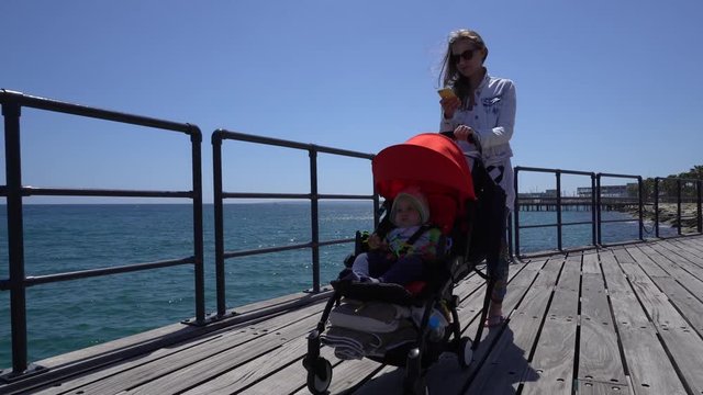 Attractive lady pushes the baby carriage on a wooden pier and messages by smartphone. Camera moves before mother, handheld shot