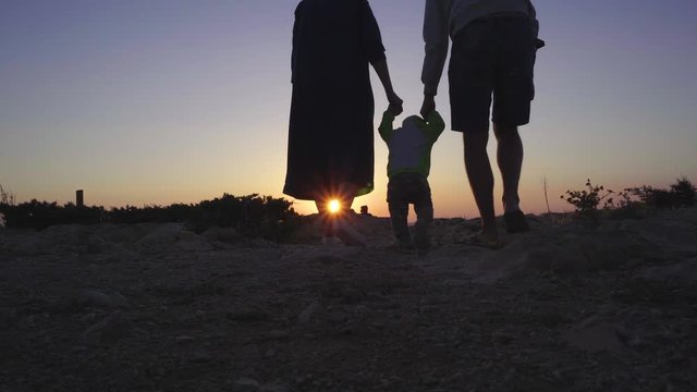 Toddler boy learns to walk, parents hald his hands and help. Young family at sunset