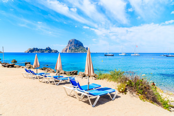 Sunbeds with umbrellas on Cala d'Hort beach with beautiful azure blue sea water and Es Vedra island in distance, Ibiza island, Spain