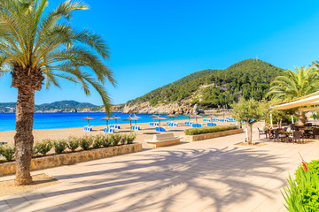 Coastal promenade along sandy beach with restaurants and bars in Cala San Vicente bay on sunny summer day, Ibiza island, Spain