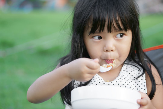 Asian children cute or kid girl and disheveled hair with hungry eating instant noodle delicious on nature camping trip, soft focus