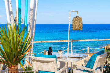 Fototapeta na wymiar White table and chairs with blue pillows on restaurant terrace on Cala Nova beach with beautiful sea view, Ibiza island, Spain