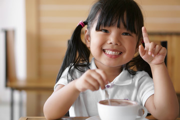 Asian children cute or kid girl white teeth smile and hot cocoa or chocolate drinking for breakfast and want one more because delicious in the morning on wood table in the home or cafe