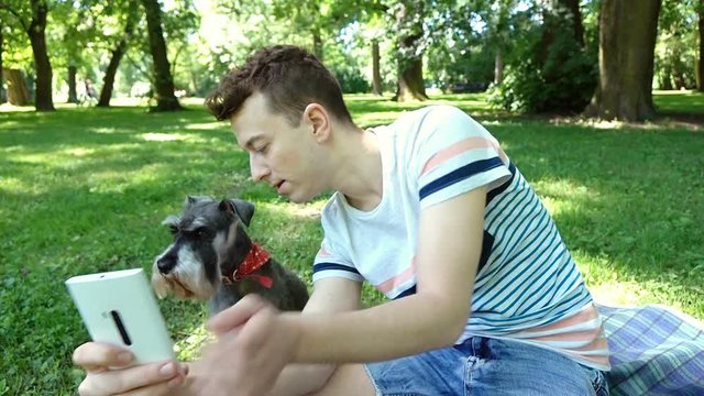 Boy relaxing with his dog in the park and doing selfies on smartphone, steadycam shot
