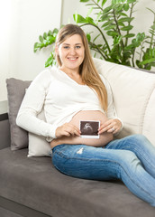 Smiling pregnant woman sitting on sofa and holding ultrasound fetus scan