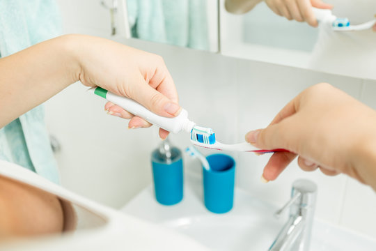 Closeup image of woman putting toothpaste on toothbrush in cup at bathroom