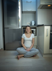 Woman in pajamas sitting on floor at kitchen next to open refrigerator at late evening
