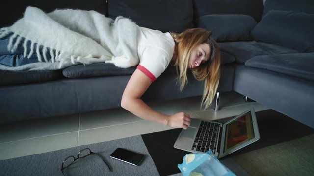 Young lazy cute female with blonde hair lies on grey sofa at home, scrolls social networks on her laptop hanging down on floor and eats potato chips.
