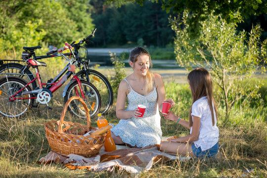 Woman Having Picnic By The River With 10 Years Old Daughter. Two Bicycles On The Background