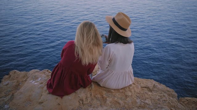 Camera moves toward two girl friends sitting on the edge of rock cliff next to ocean, taking photos on small digital camera and talking about something