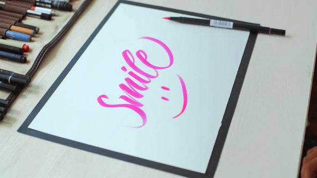 Handheld shot on illustrator hands drawing nice smile icon below his calligraphy Smile on white a4 paper sheet on wooden desktop