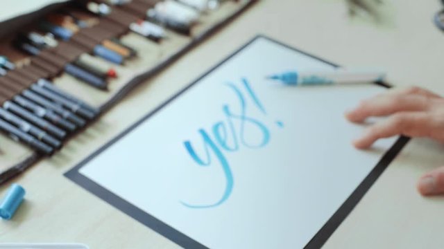 Handheld shot of artists hands write word yes with positive blue color on white a4 paper sheet on wooden desktop, close up shot
