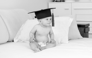 Monochrome funny image of naked baby in diapers and graduation cap sitting on sofa