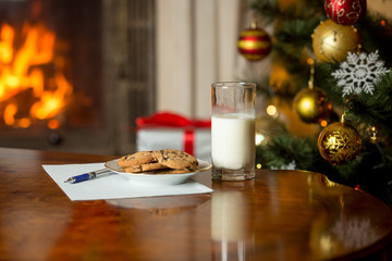 Treats and letter to Santa on wooden table next Christmas tree and burning fireplace