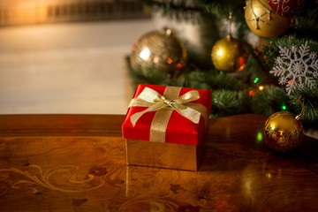 Christmas background with red gift box on wooden table in front of burning fireplace and Christmas tree