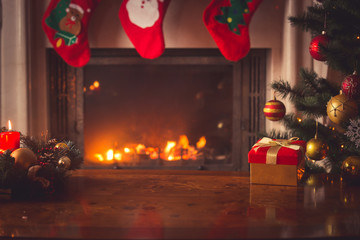 Closeup of red Christmas gift box with golden ribbon next to decorated Christmas tree and fireplace