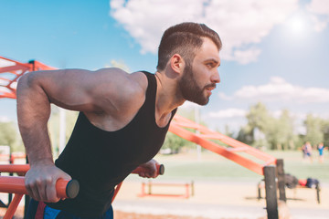 A portrait of a focused muscular bearded man in black workout clothes doing dips on parallel bars . Mans fitness with blue sky in the background and open space around him. Sports and crossfit