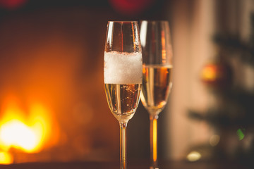 Person filling two glasses with champagne. Burning fireplace and decorated Christmas tree