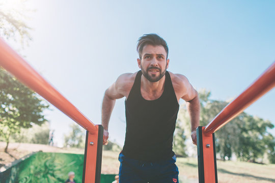 A portrait of a focused muscular bearded man in black workout clothes doing dips on parallel bars . Mans fitness with blue sky in the background and open space around him. Sports and crossfit