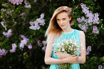 Blonde girl with bouquet of wild flowers outdoors A model in a turquoise dress against a background of a lilac bush.