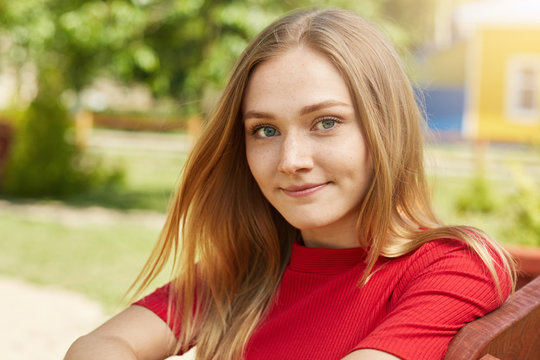 Headshot of beautiful blonde girl having warm eyes, freckles and sincere smile looking pleased into camera while having rest at bench in garden. People, facial expressions and lifestyle concept