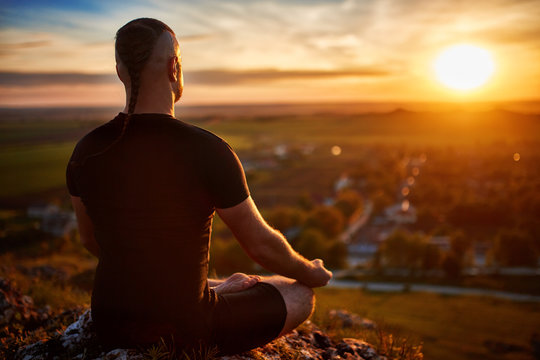 Rear view of the man meditating yoga in lotus pose on the rock at sunset.