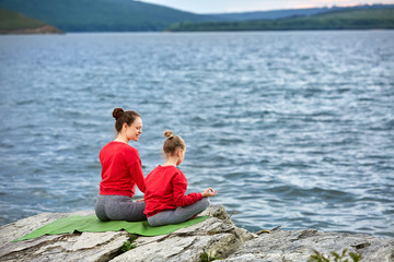 Rear view of the mother and daughter are practicing yoga on the rock near river.