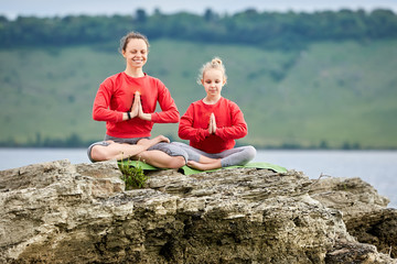 Sporty mother and daughter doing yoga on the rock near beautiful river.