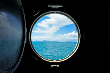 Seaview with blue sky and white clouds from outside of vintage circle porthole or window frame at rusted steel side wall on old ferry boat.