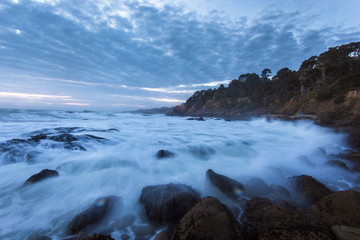 Obraz premium Dramatic ocean tide splashing water by rocky shore during sunset in California
