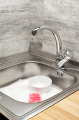 Kitchen sink full of foam, tableware and pink cleaning sponge