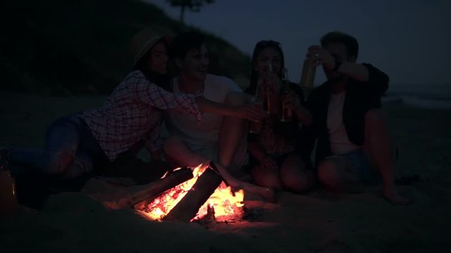 Picnic of young people with bonfire on the beach in the evening drinking beer and doing cheers. Happy friends singing songs and playing guitar. Slowmotion shot