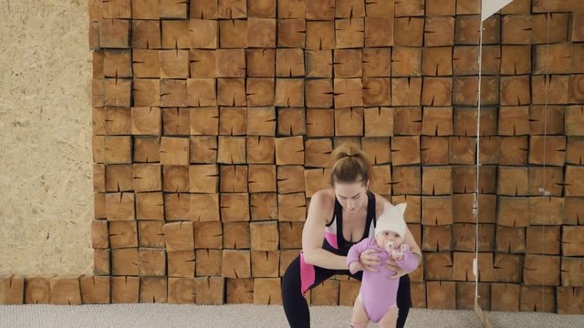 Attractive woman doing exercises holding little child indoors. Standing straight with widely spaced legs she slowly squat down to parallel with floor, lowering baby and returning to starting position