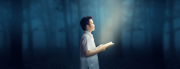 Young School Boy in uniform holding a book reading in the dark cold forest at night.