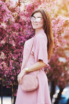 Outdoor portrait of young beautiful fashionable elegant girl with long hair posing in street. Model wearing stylish pink dress, sunglasses, carry small bag. City lifestyle, female fashion concept