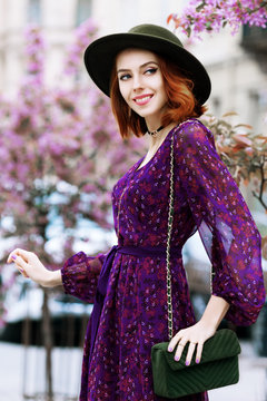 Outdoor portrait of young beautiful happy smiling woman posing in street. Model wearing stylish purple dress, green fedora hat, carry small shoulder suede bag. City lifestyle. Female fashion concept