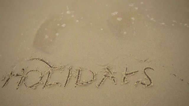 Summer vacation concept. The word holidays written in the sand on beach, waves washing it aways.