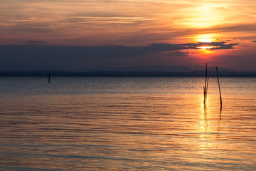 Beautiful sunset with sun behind some clouds and reflecting on a lake, with red and orange tones in the sky and clouds