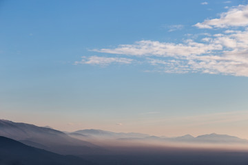 Plakat View of a valley at golden hour, with mist between the hills and mountains, and a blue sky with white clouds