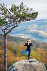 Naklejka na ściany i meble Peaceful fit female is practicing yoga and doing asana Vrikshasana on the top of the mountain near big tree. Autumn forests and hills on the background