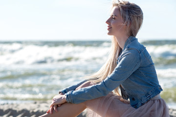 Fototapeta na wymiar Young cheerful girl on the seashore. Young blonde woman sitting on the sand. Trendy beige skirt