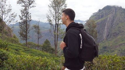 Young man tourist with backpack walking at trail in mountains with beautiful nature landscape at background. Male hiker going along tropical mount road. Healthy active lifestyle. Travel concept