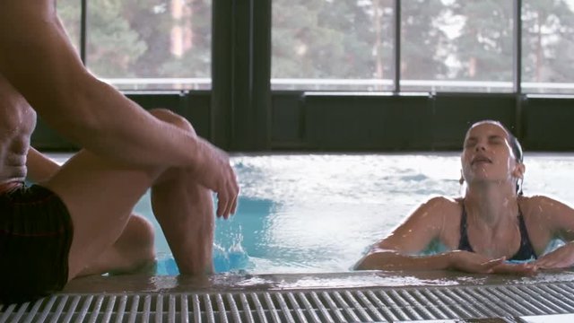 PAN with slow motion of happy young woman diving into indoor swimming pool with blue water, then emerging and chatting with muscular middle-aged man 