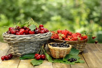 Fototapeta na wymiar Summer gifts: sweet cherry, strawberry and black currant in wicker baskets on wooden desk on background of green leaves